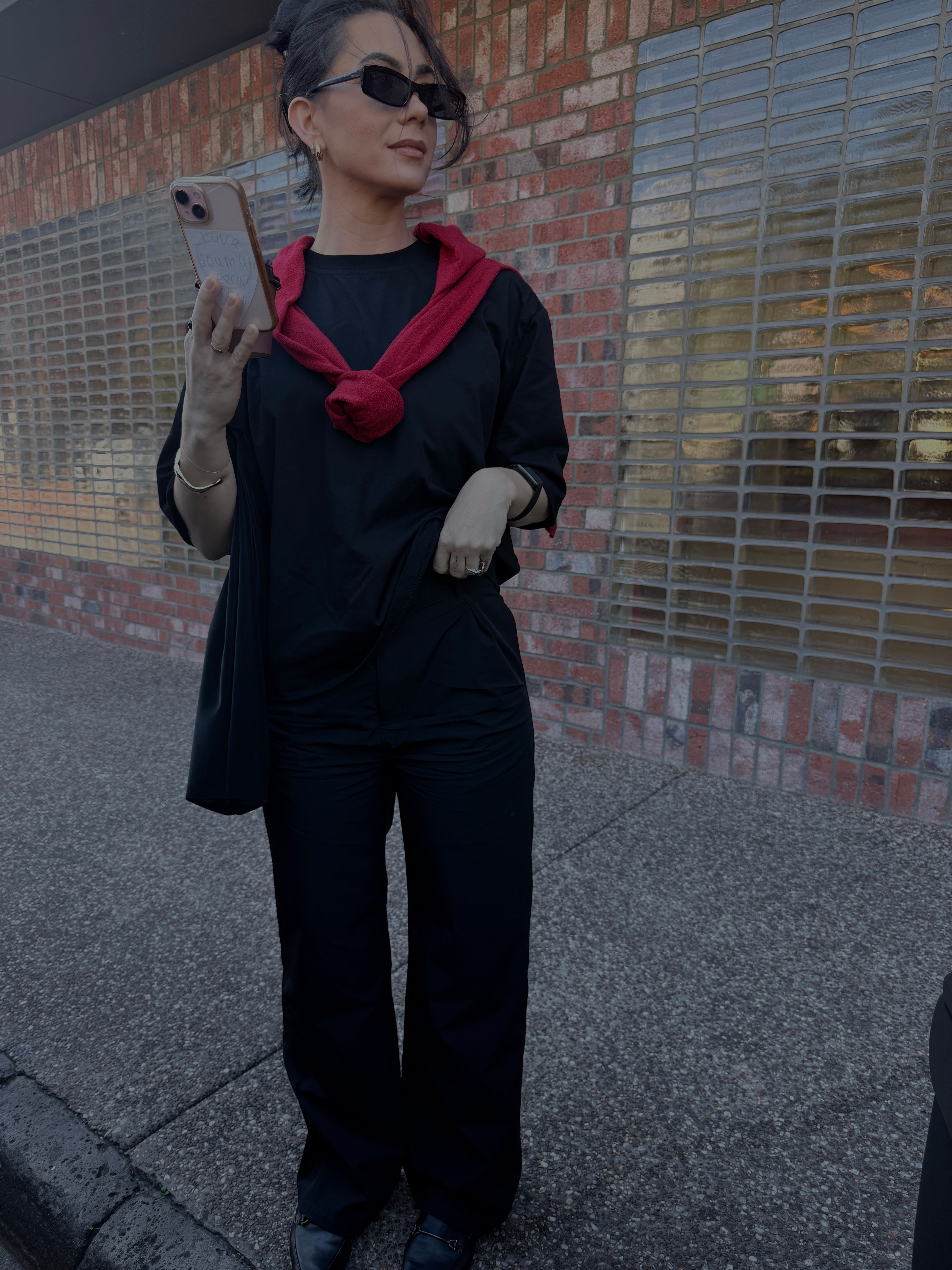Person wearing the black drew pants and black shoulder pad tshirt with a red scarf, standing on a sidewalk.