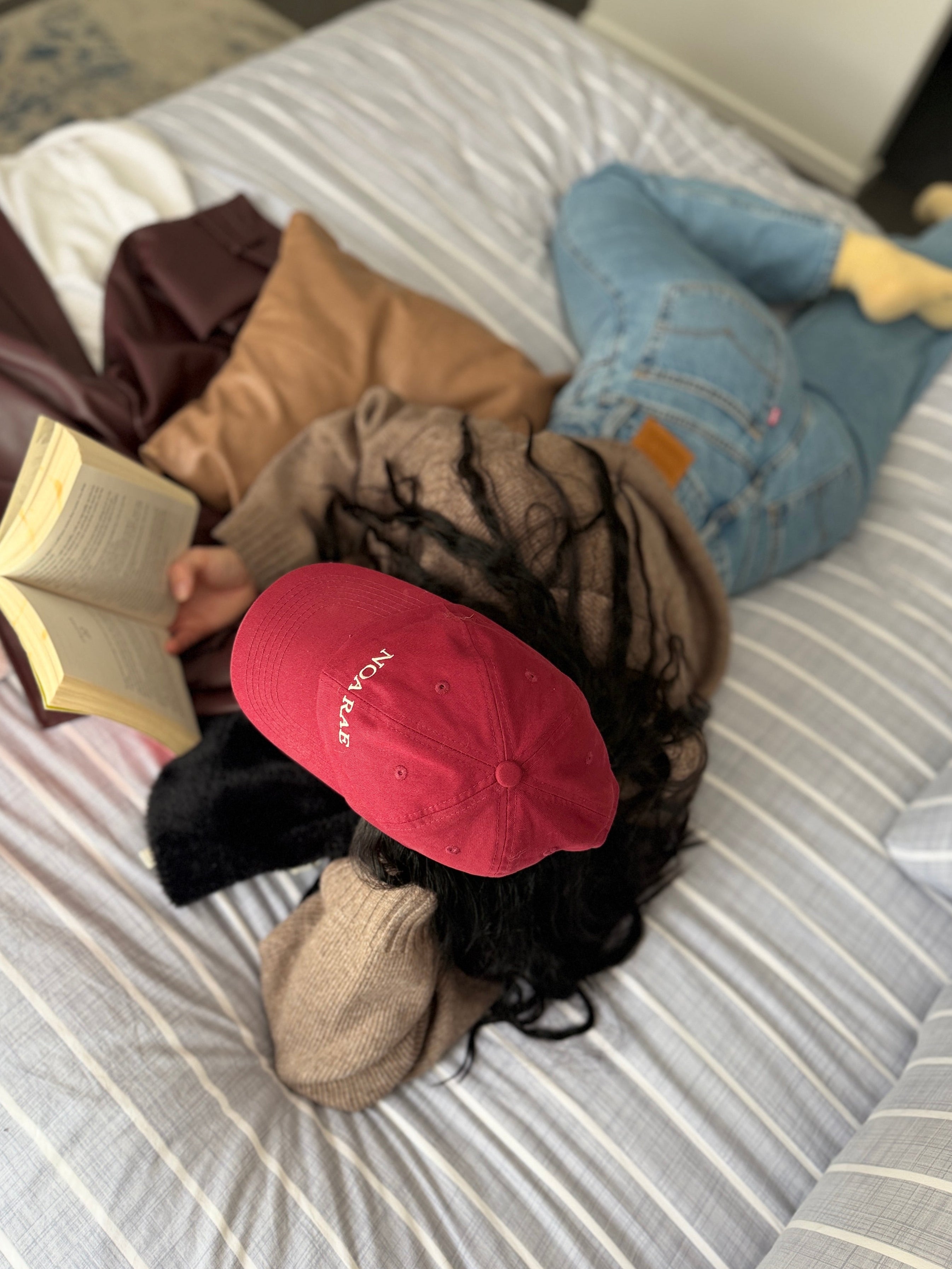 Person wearing a red/Plum Noa Rae cap and reading a book on a bed